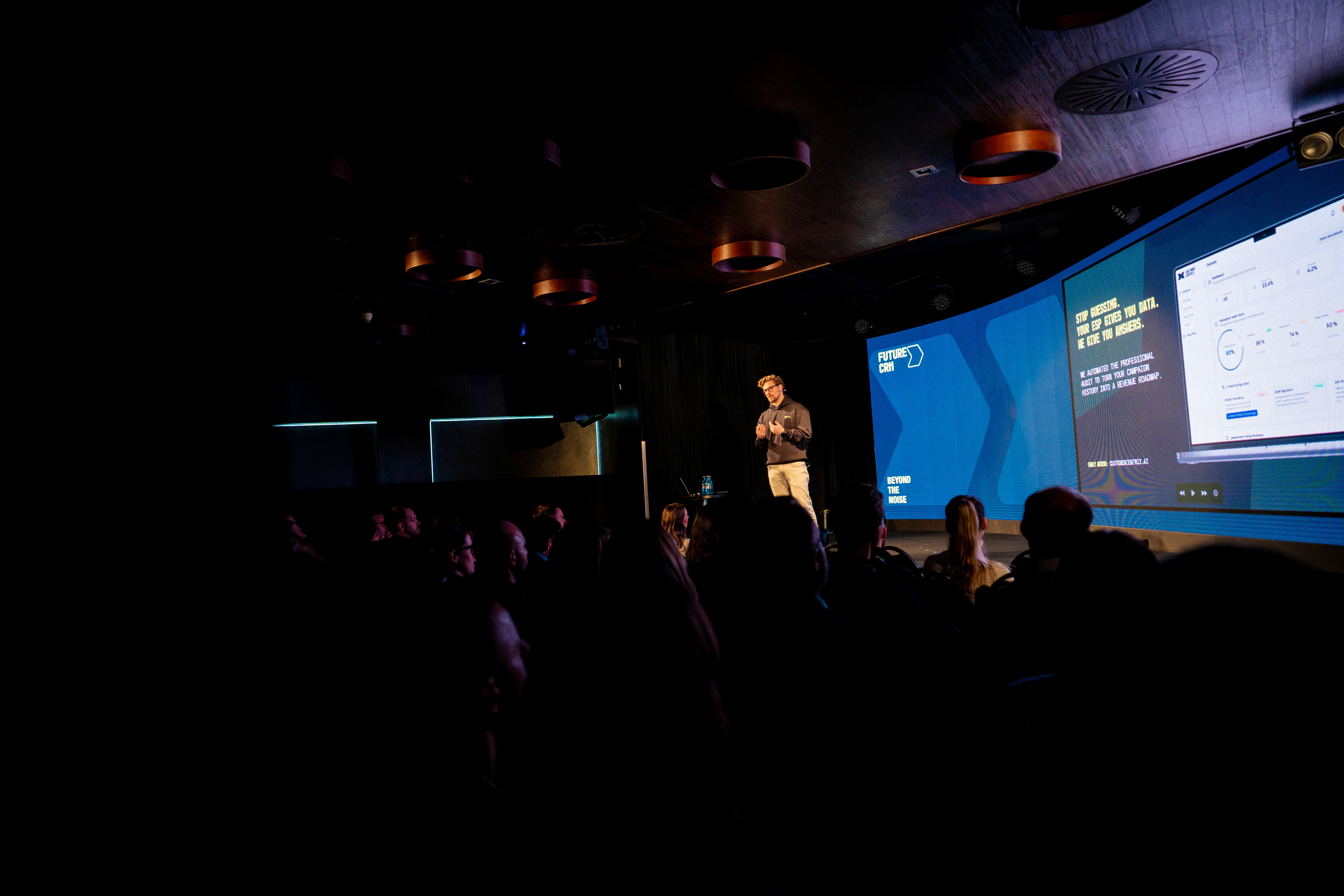 A speaker stands on a stage presenting to an audience in a dark room, with a large screen displaying graphs and text beside him. The audience is seated and mostly in shadow.