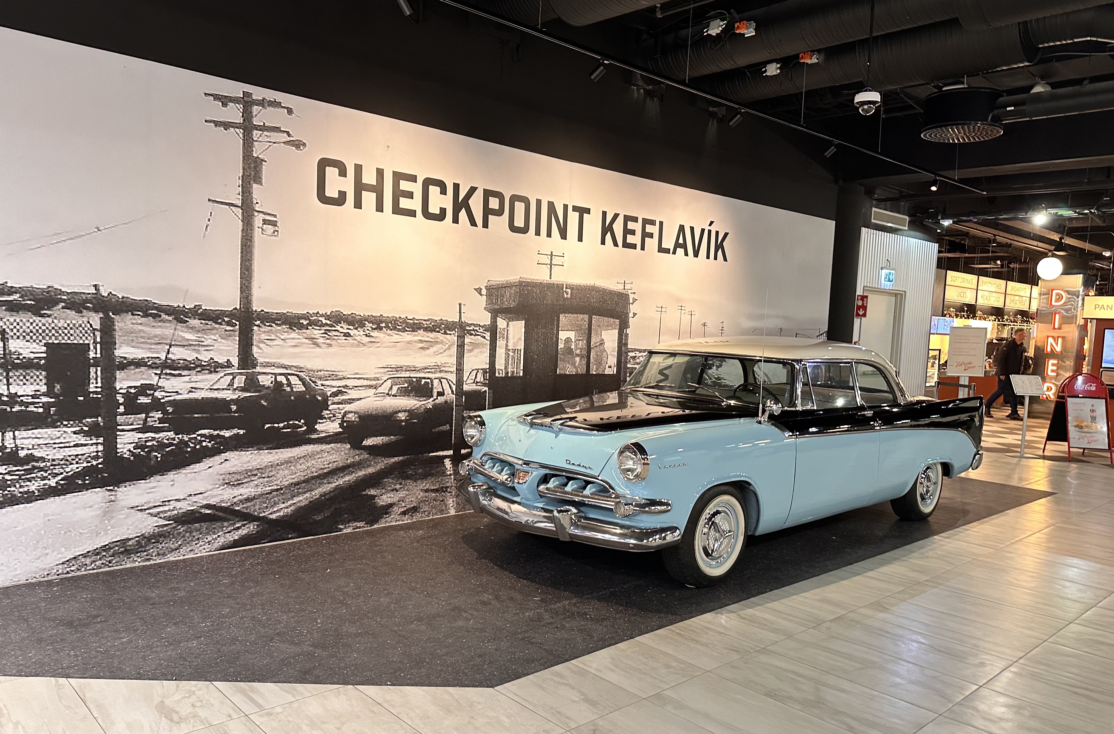 A vintage light blue and black car is displayed indoors in front of a large mural that reads "CHECKPOINT KEFLAVÍK." The mural depicts a historical checkpoint scene with cars, a guard booth, and fencing, evoking Cold War-era imagery. The display appears to be part of a museum or exhibition space.