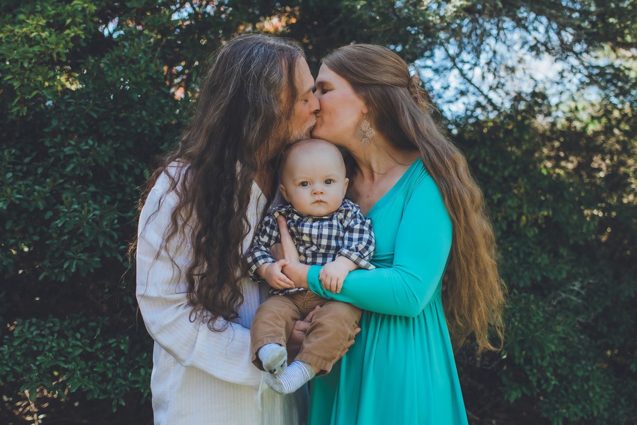 Two women kissing with a baby between them. Family photo with the women wearing white and teal dresses holding the baby.