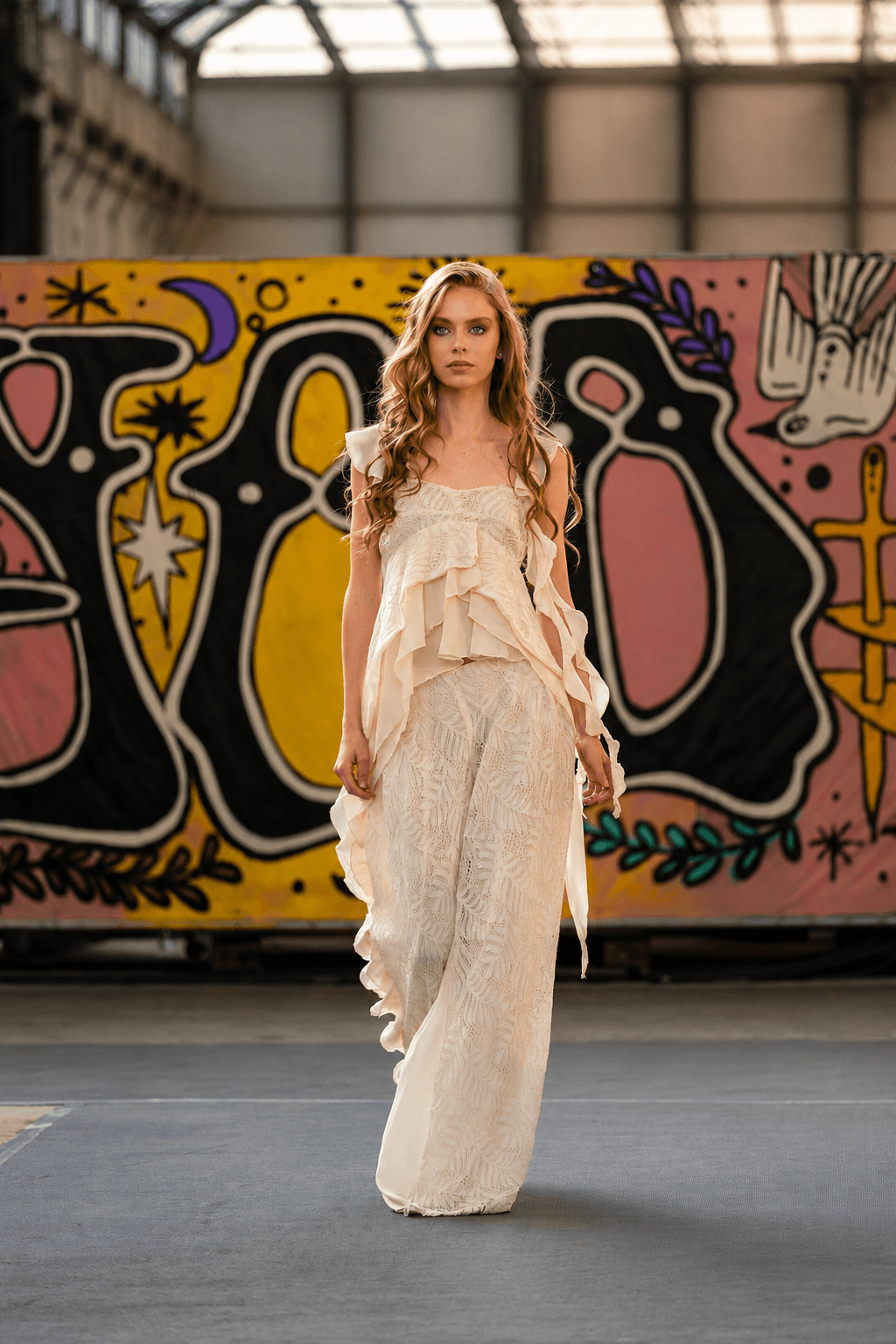 Nelly Opitz walks the runway in a flowing cream outfit against a colorful graphic backdrop.
