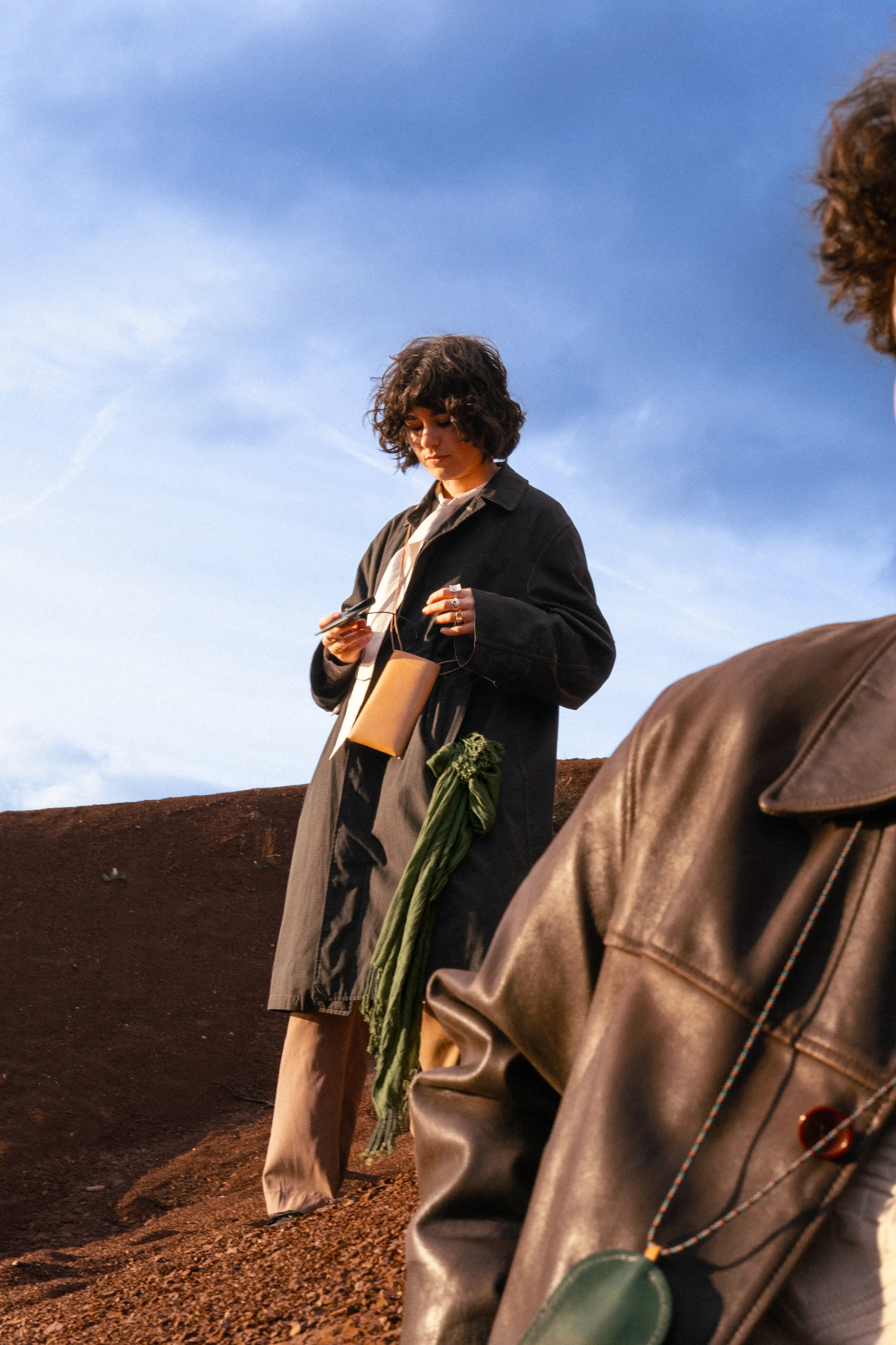 Anaïs Brancheriau standing on red earth under a blue sky, wearing a long dark coat, beige trousers, and a small tan crossbody pouch, with a green scarf hanging from one hand and a blurred leather-clad figure in the foreground.