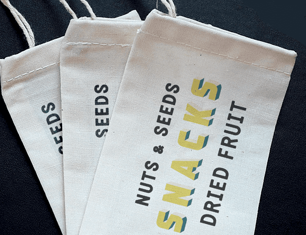 Three off-white fabric bags with stitching and drawstrings are stacked on a dark fabric surface. The top bag displays the text "NUTS & SEEDS" in black letters, with "SNACKS" and "DRIED FRUIT" in yellow-green letters below. The middle bag shows "SEEDS" in black, and the bottom bag also displays "SEEDS" in black.