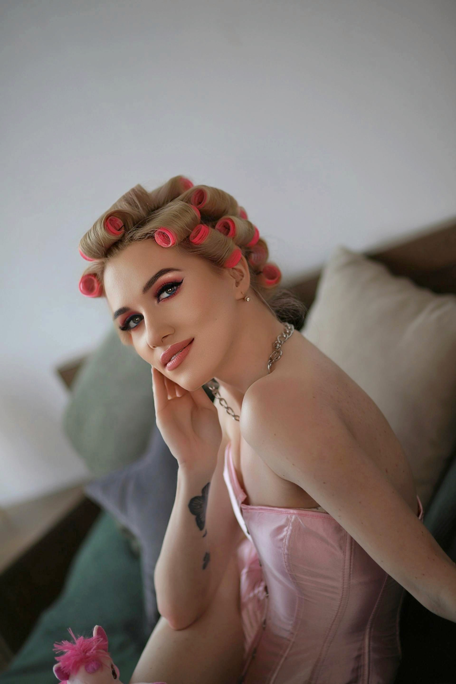 A woman with blonde hair in pink rollers and a pink corset sitting on a bed, resting her face on her hand and looking toward the camera.