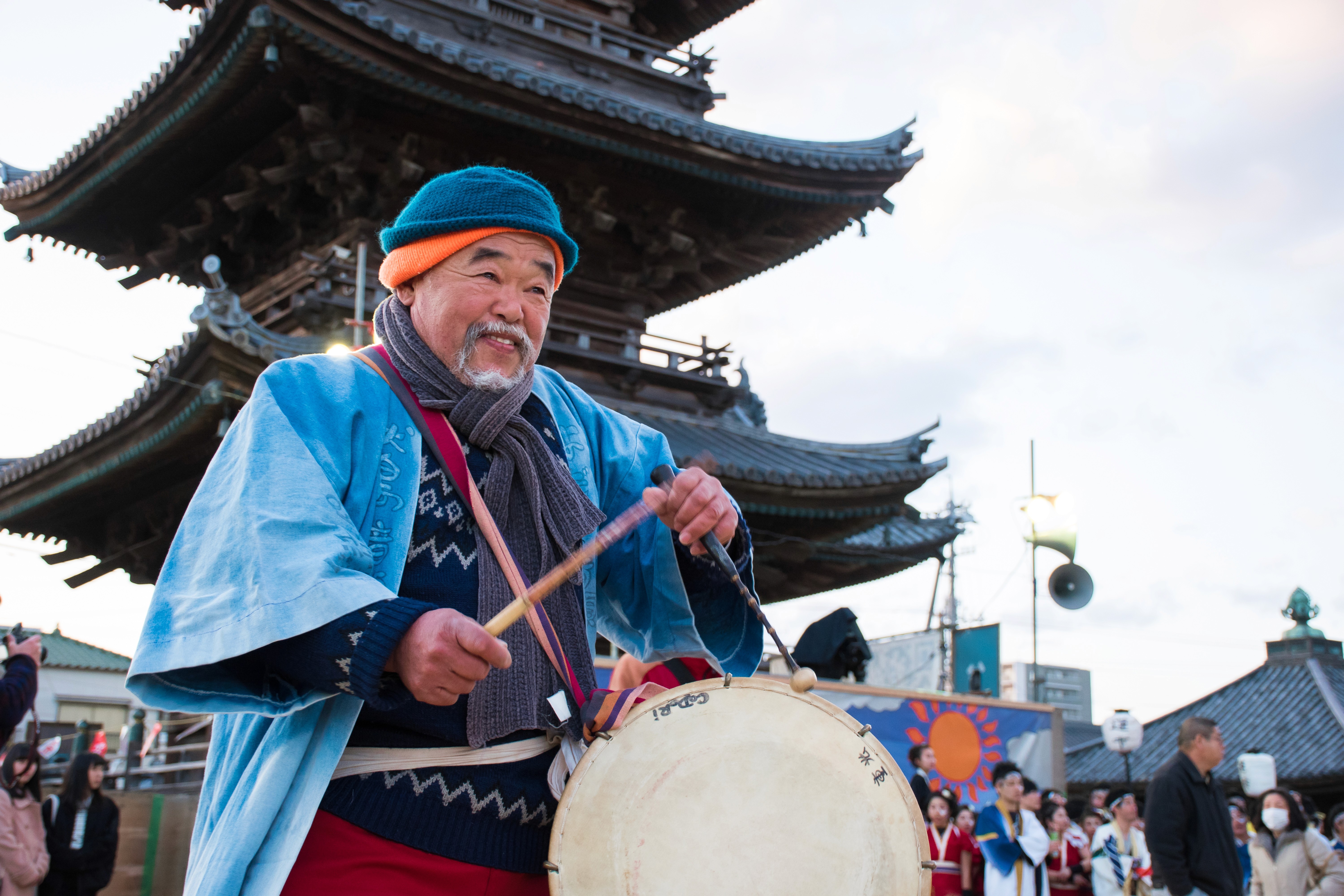 Older Japanese man wearing festival clothes, playing a traditional drum in front of a pagoda