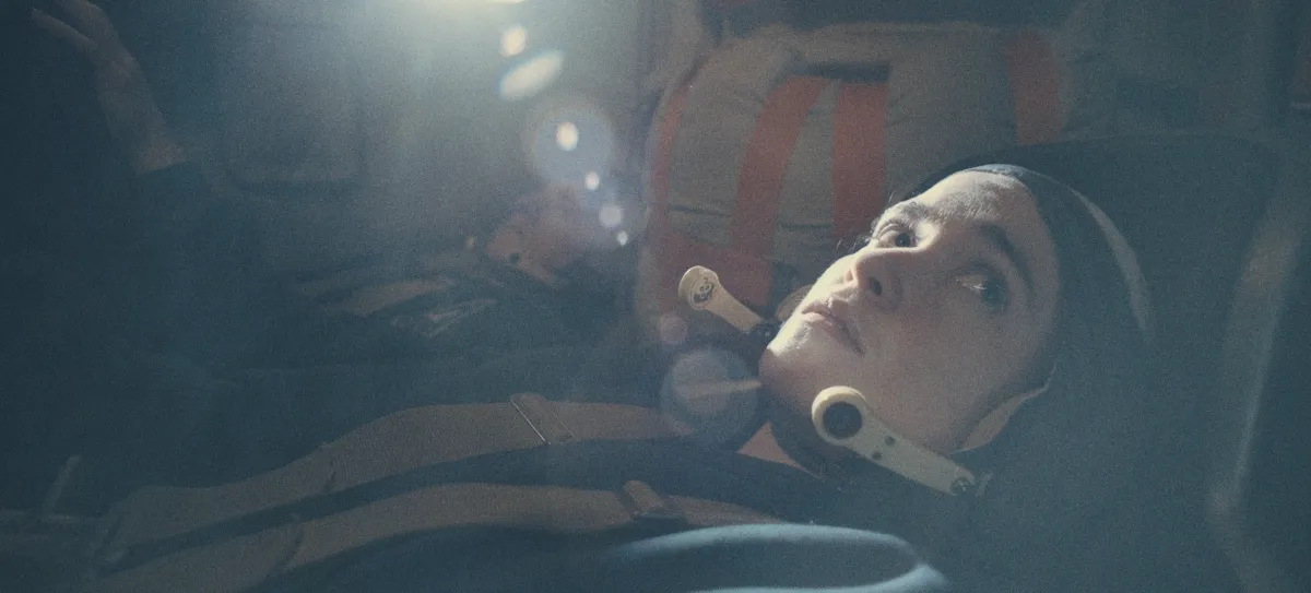 A young woman lies in a dimly lit interior, wearing a helmet and harness while illuminated by a soft, ethereal light, suggesting a scene from a space mission or simulation.