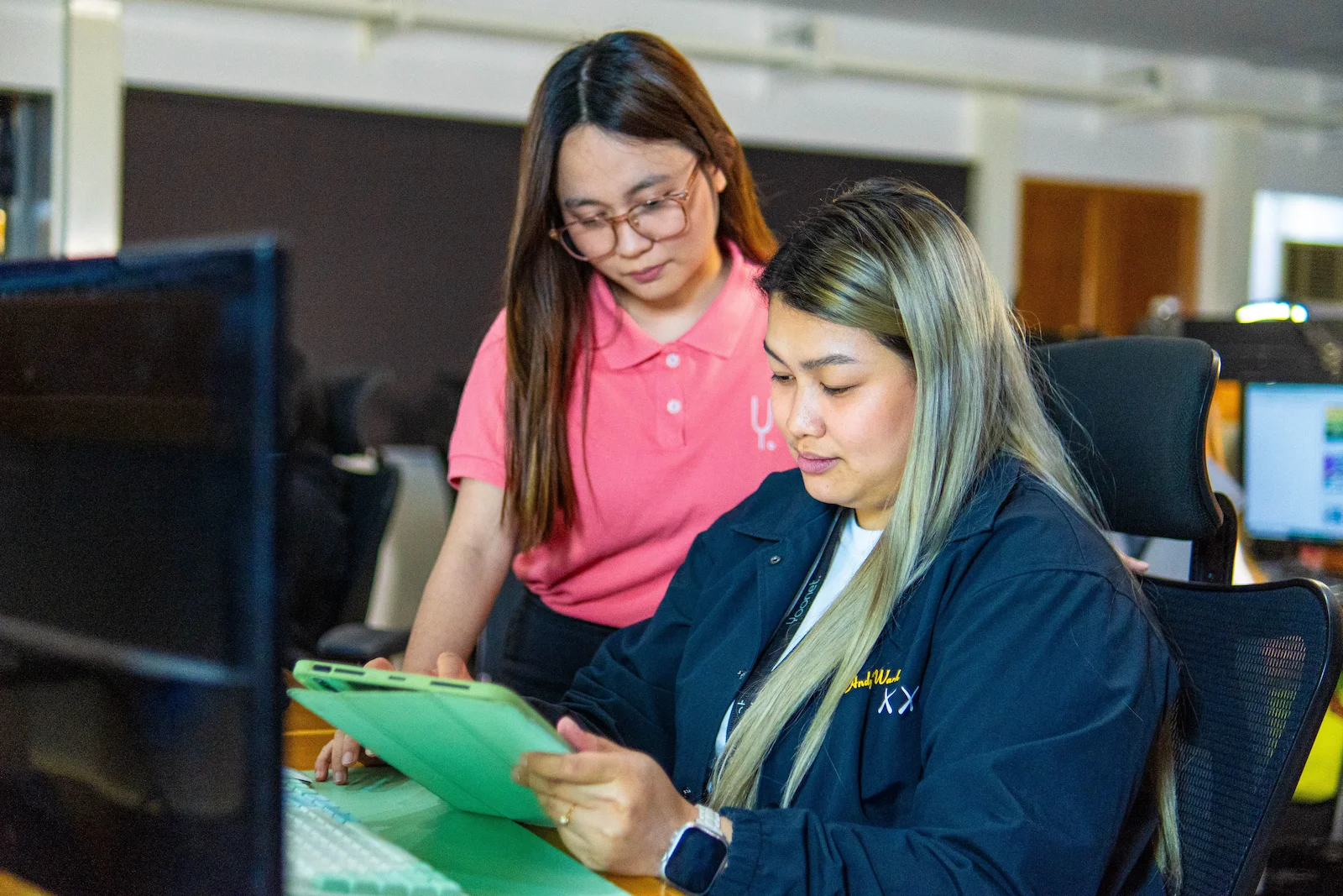 Two women work together in an office setting, one standing and pointing at a document while the other sits at a computer.