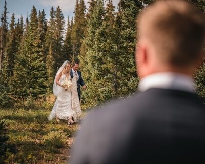 Seen over the groom's shoulder, the bride walks toward the ceremony with an escort.