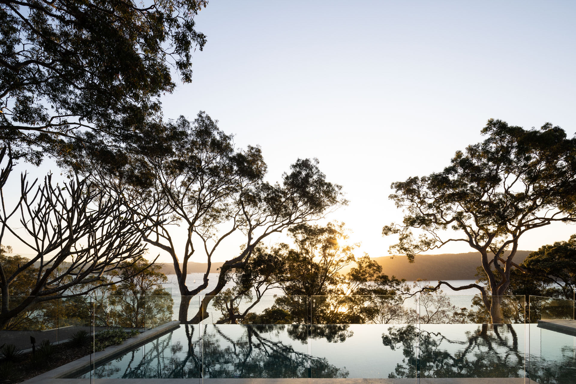 A spacious industrial-style interior with exposed brick walls, large windows, and natural light pouring in during sunset. Wooden desks with plants and workspace materials are arranged along the walls, with a view of greenery and an outdoor seating area.