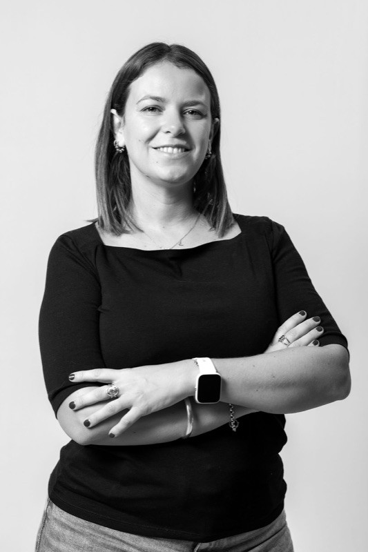Black-and-white studio portrait of a smiling woman with straight shoulder-length hair, wearing earrings, a necklace, rings, and a black square-neck top, arms crossed, against a plain light background.
