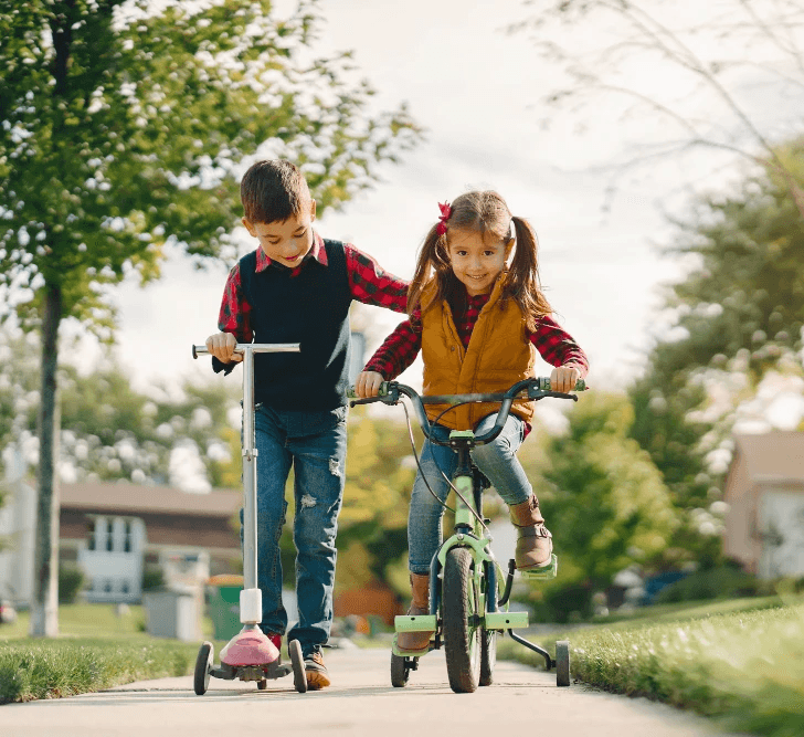A young boy and girl play outside on a sunny day, riding a scooter and a bike along a suburban sidewalk. The image represents the freedom and family-friendly lifestyle made possible through USDA Rural Development home loans from Chris Lewis Home Loans.