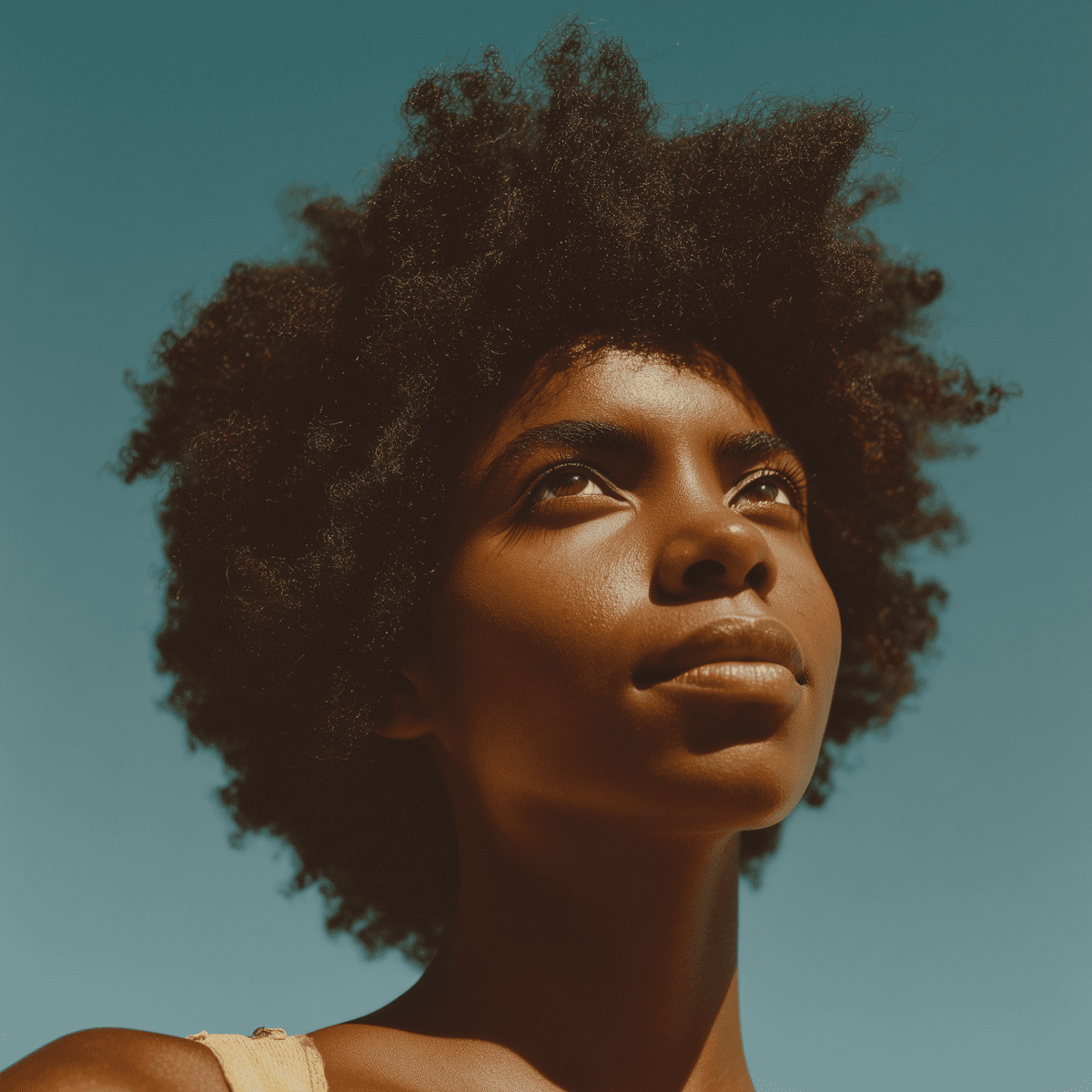Low-angle portrait of a woman with an afro looking toward the sky against a clear blue background.