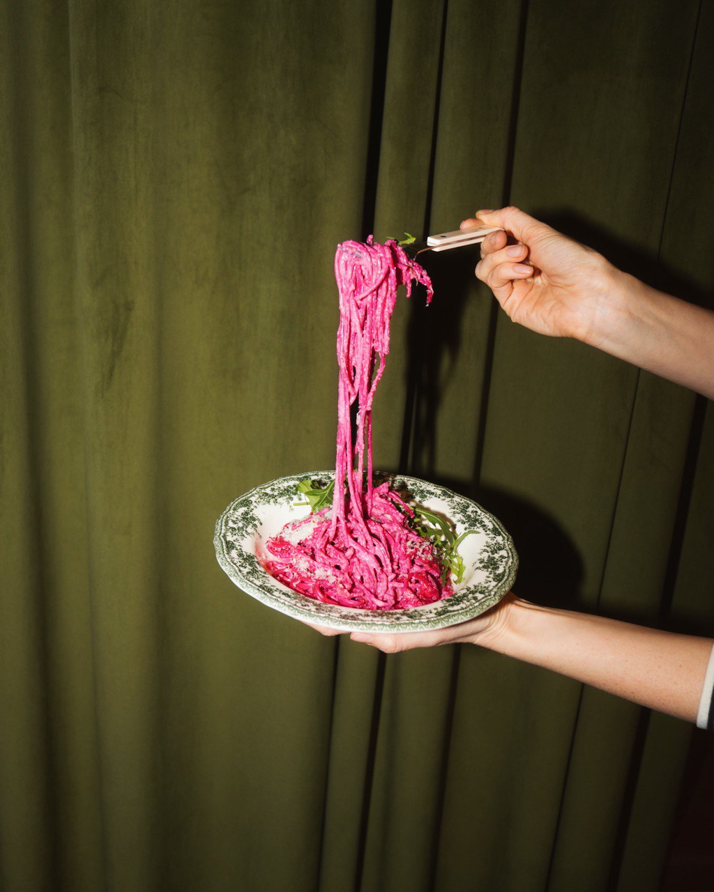 A hand holds a tall, pink dessert on a plate, with a green background behind it.