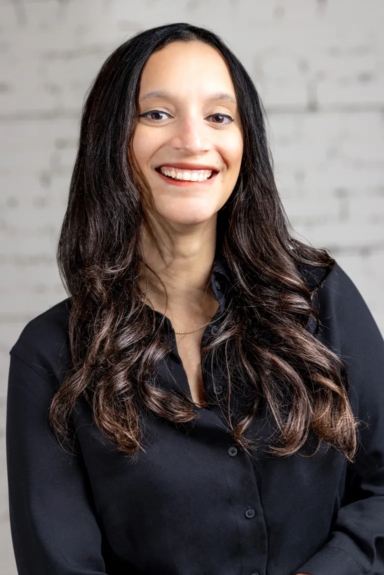 Image of Smiling woman with long dark hair wearing a black shirt standing in front of a wall.