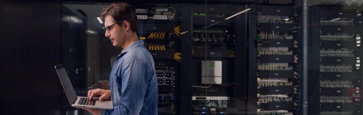 Ein Techniker, der in einem Serverraum an einem Laptop arbeitet, während im Hintergrund Netzwerkkabel und Server zu sehen sind, betont die Operationalisierung der Sicherheit und die ununterbrochenen SecOps.