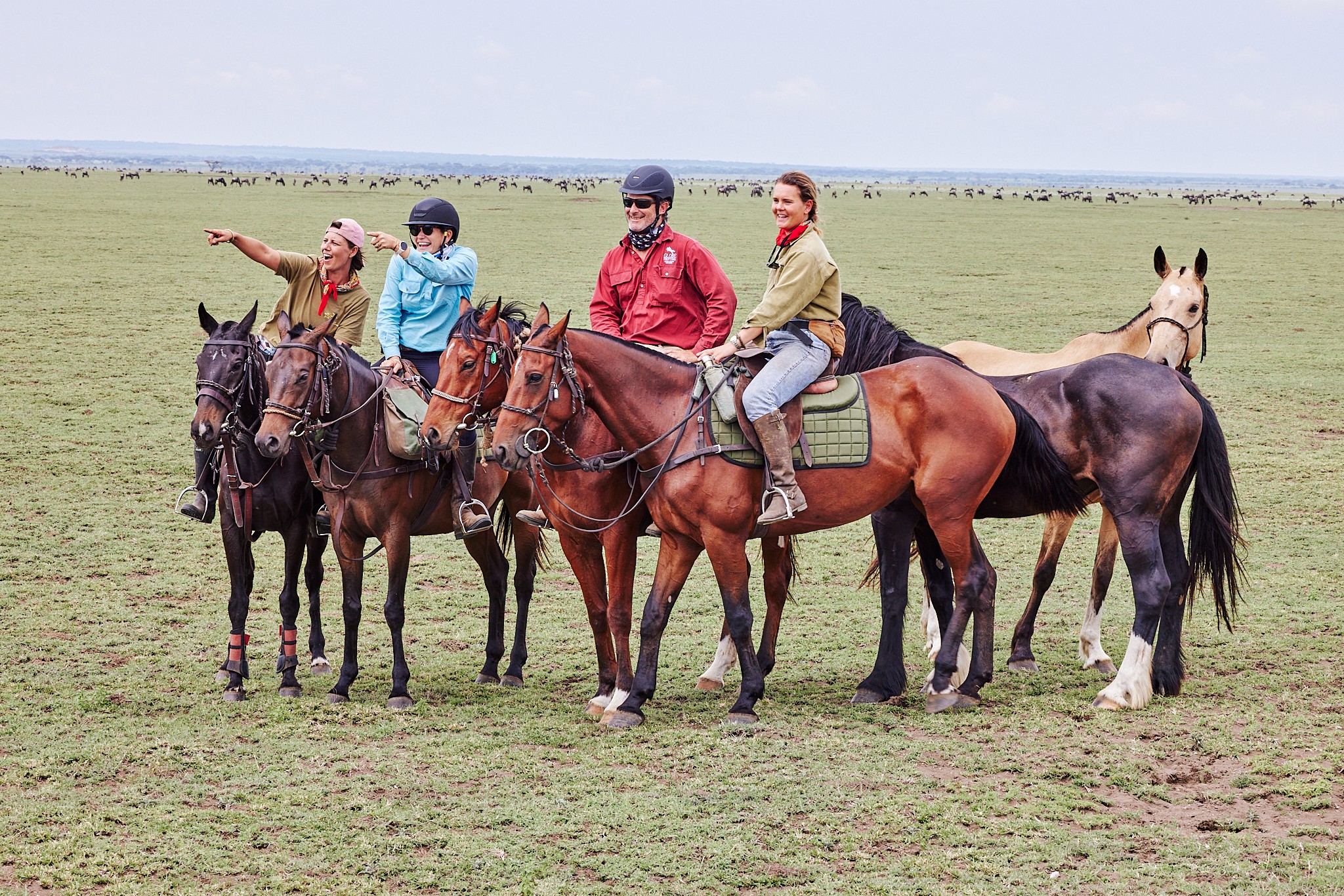 Grupp av ryttare på ridresa i Afrika galopperar över Serengetis vidsträckta slätter medan damm och gnuhjordar rör sig över horisonten under the great migration.