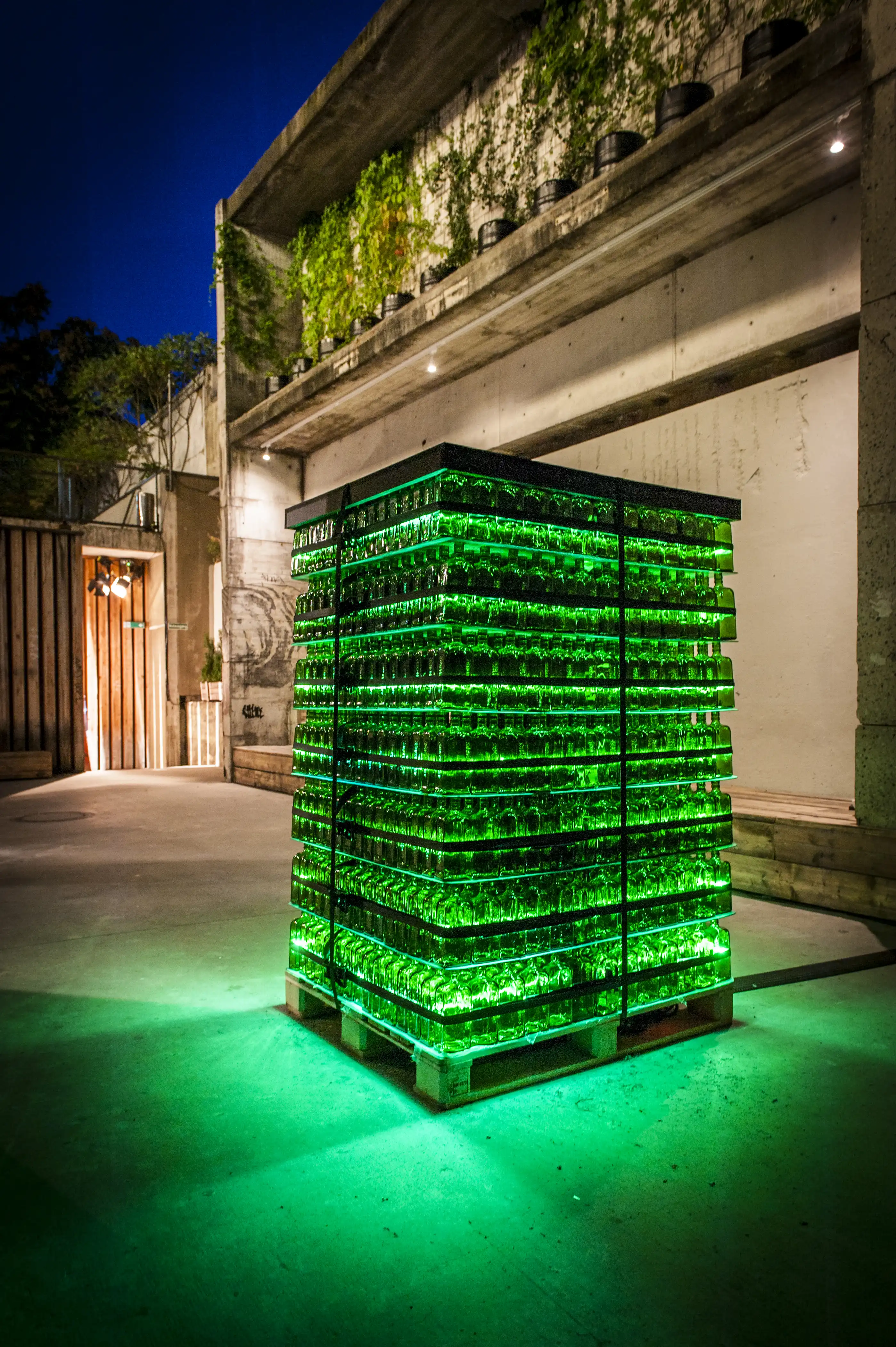 Green-lit tower of stacked Jägermeister bottle crates on a pallet in an industrial concrete courtyard at night.