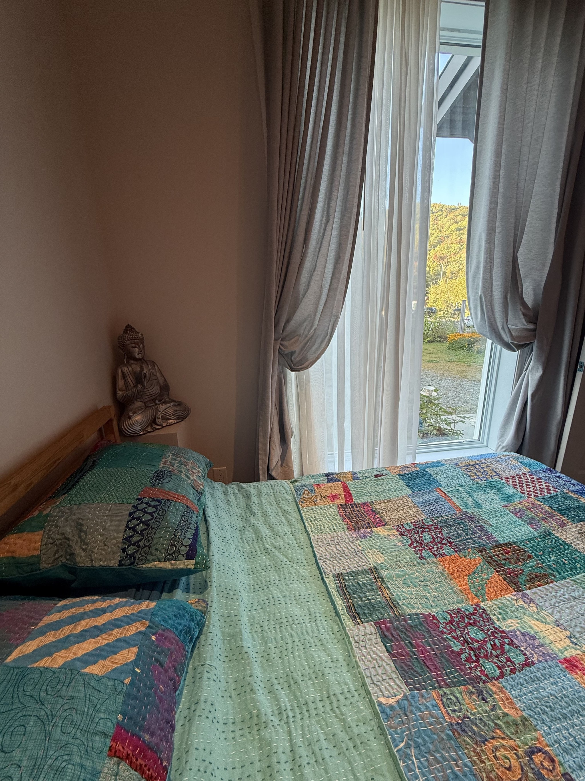 Lit double aux couvertures patchwork colorées, statue de Bouddha et grande fenêtre donnant sur la nature.