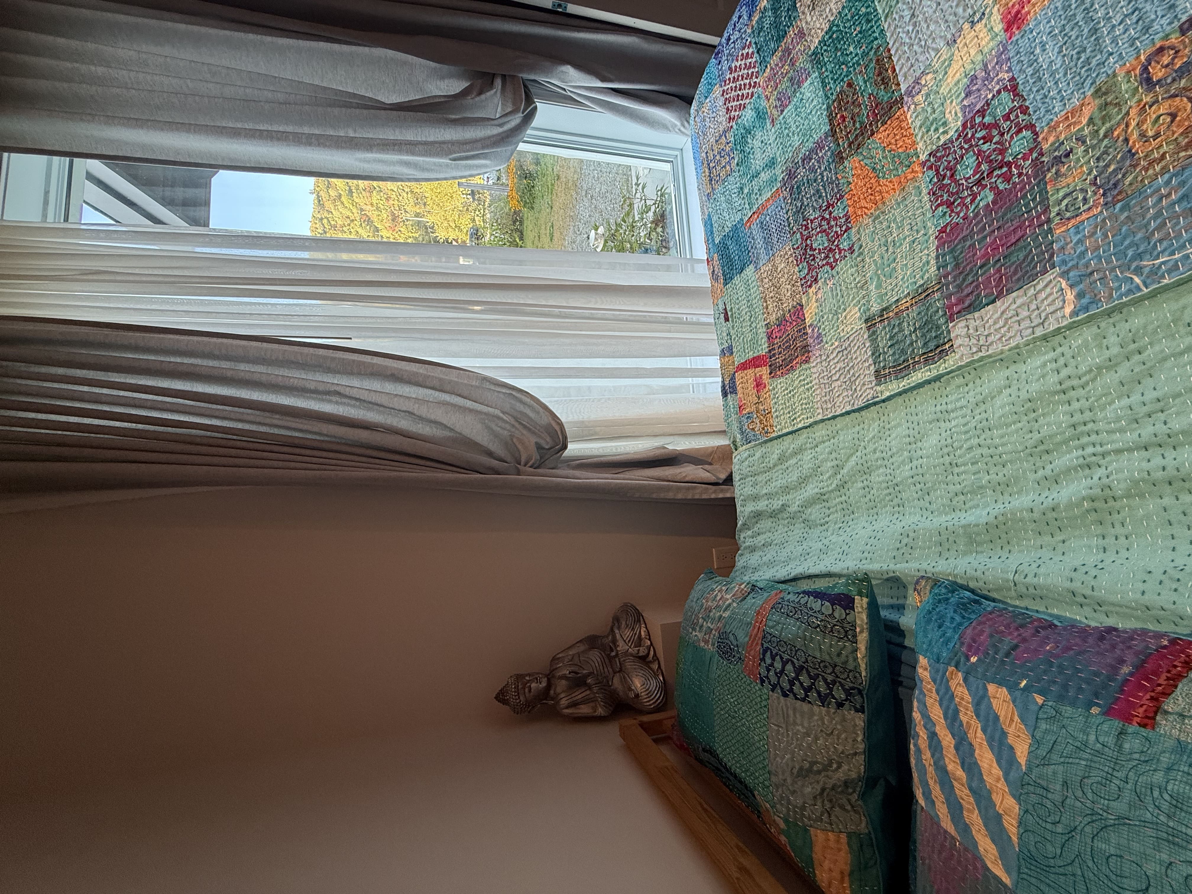 Lit double aux couvertures patchwork colorées, statue de Bouddha et grande fenêtre donnant sur la nature.