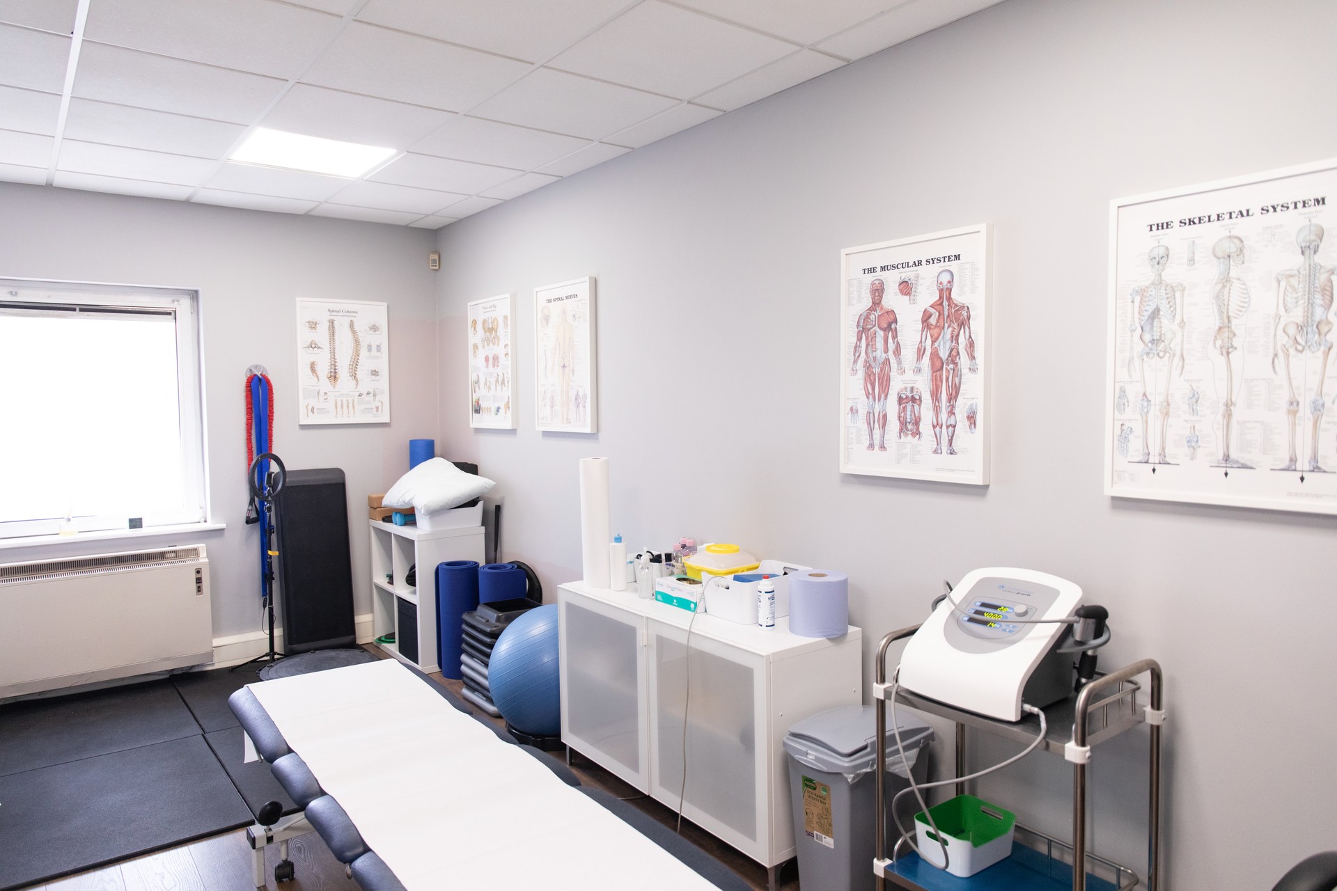 A treatment room with a treatment couch, a physio ball, a shelving unit and a treatment machine, with anatomy charts on the walls.