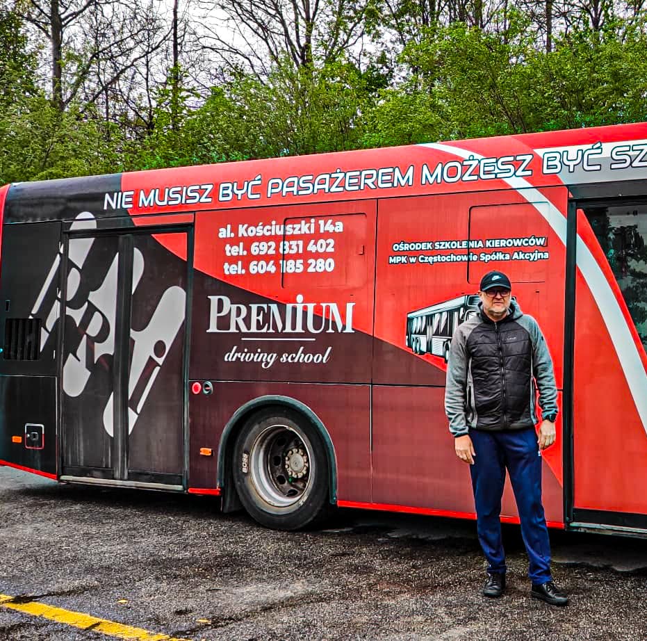 Maciek – Instruktor oparty o srebrny pojazd nauki jazdy z logiem Premium w słoneczny dzień.
