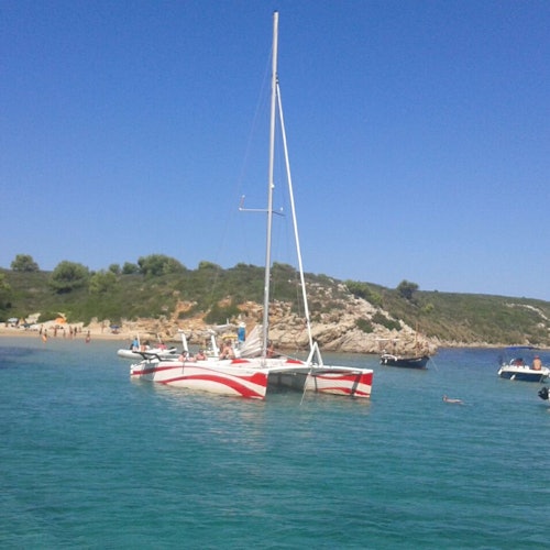 En röd och vit katamaran flyter på klart, turkost vatten nära en strand med människor och en klippig kustlinje under en blå himmel.