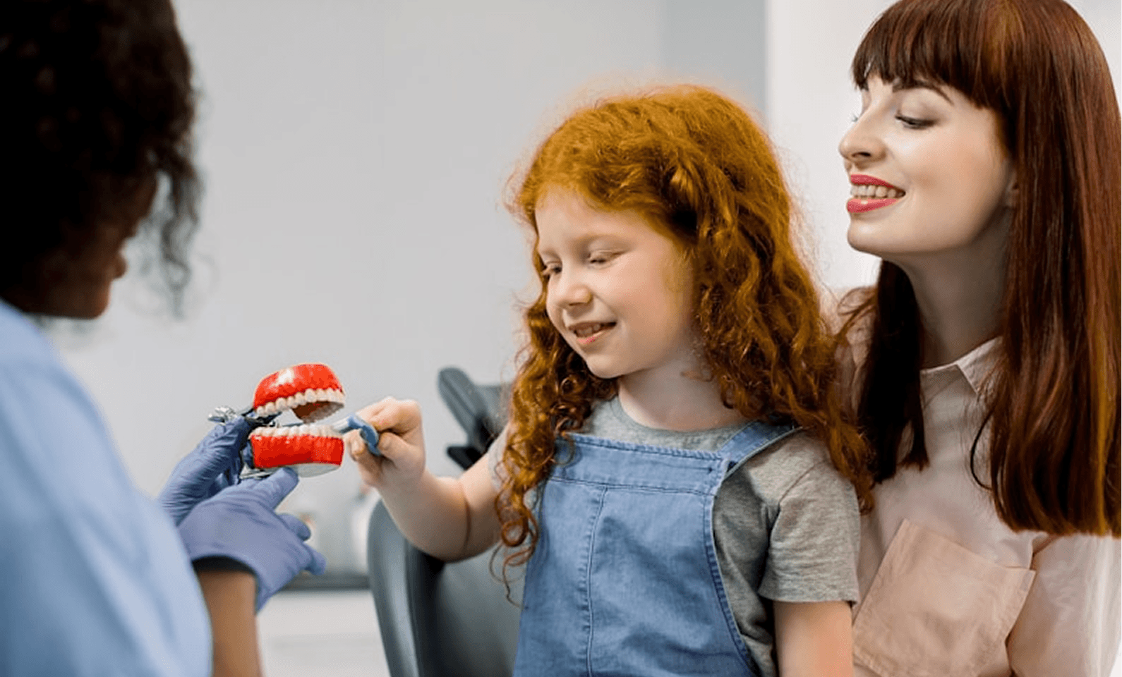 A child with curly red hair, wearing a denim overall, enthusiastically holds a dental model while sitting next to a smiling adult with straight red hair, while a professional in a dental uniform and gloves assists, highlighting child-friendly dental care in a clinic setting.