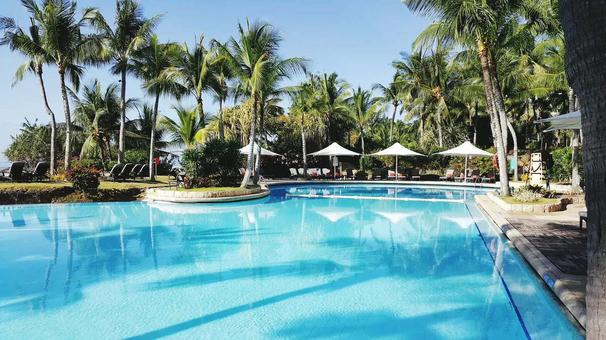 Tropical resort swimming pool surrounded by palm trees, sun loungers and white umbrellas on a bright sunny day.