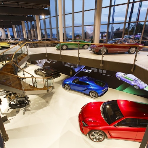 Un showroom con varios autos deportivos exhibidos en dos niveles, grandes ventanas al fondo y una escalera de caracol a la izquierda.