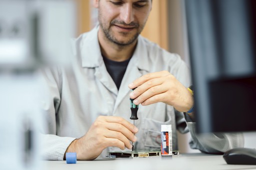 A man in a lab coat precisely working on a circuit board with a wrench at his desk, a glue stick nearby. Blurred screens and desk items are in the foreground.