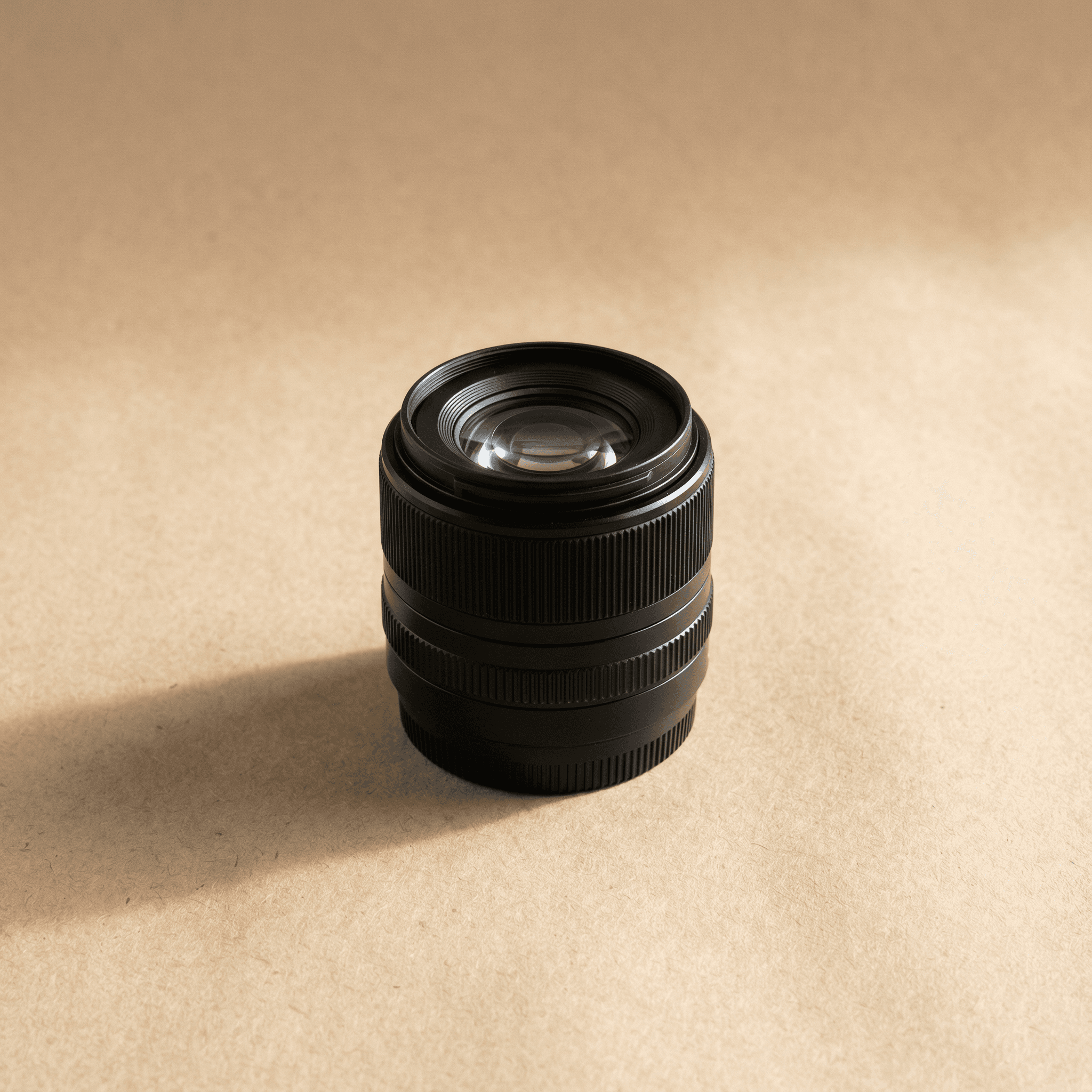 Black cylindrical camera lens on tan textured background, subtle shadows, close-up product photo of professional photography gear.