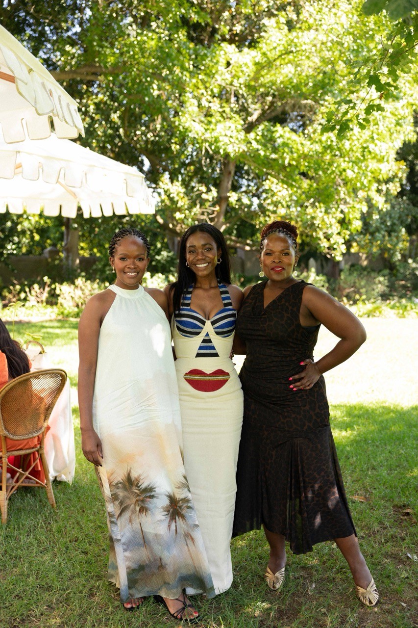 Three women, Vimbai in the centre, at The Gathering event: one wearing white dress with palm tree design, one in navy stripe dress with red lip design, and one in black dress, standing together in garden setting under white awnings