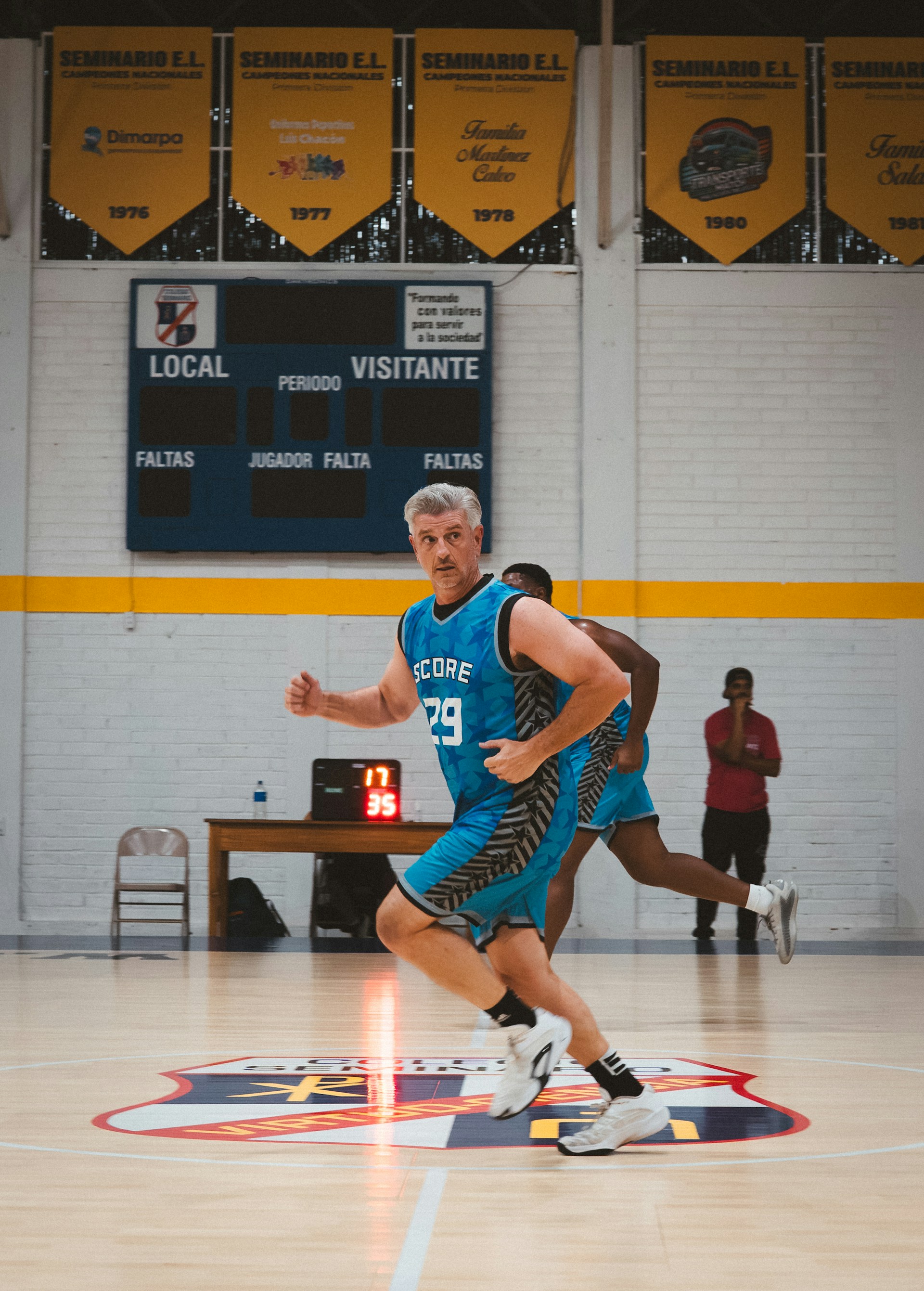 An older male basketball player with grey hair, wearing a blue jersey (number 29), running down the court during an indoor game.