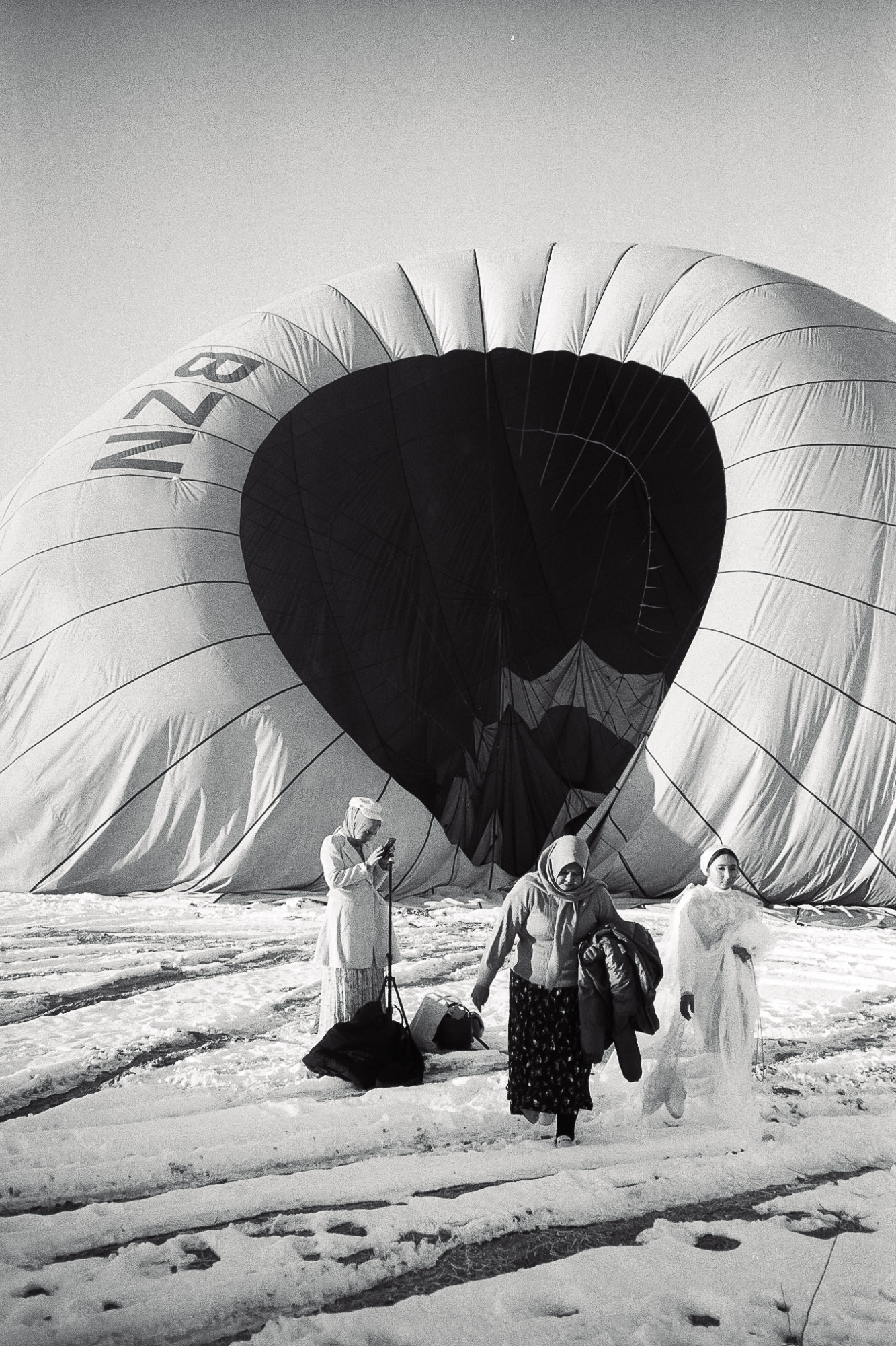 A black and white photograph showing three people dressed in winter clothing walking away from a deflated hot air balloon on a snowy field, with the balloon's fabric spread out behind them.