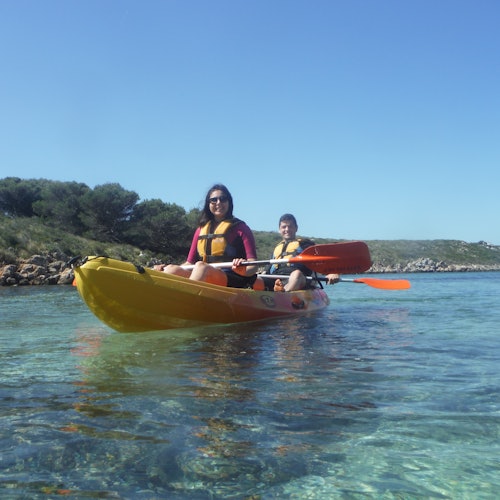 Två personer paddlar kajak på klart, lugnt vatten nära en stenig kust med träd under en blå himmel.