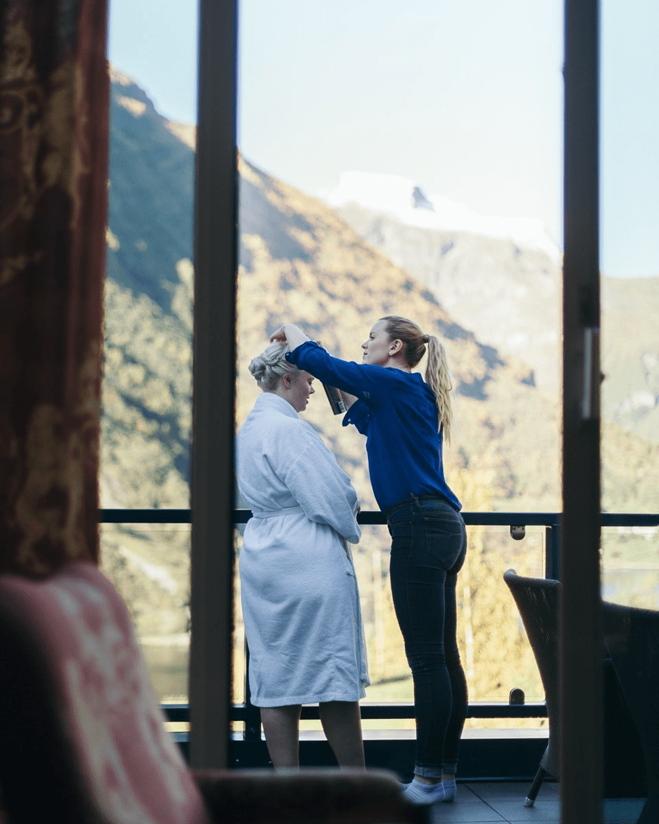 A woman in casual attire styles the hair of another woman in a white robe on a balcony, set against a backdrop of majestic mountains and a clear blue sky, suggesting a serene and intimate moment during a scenic getaway.