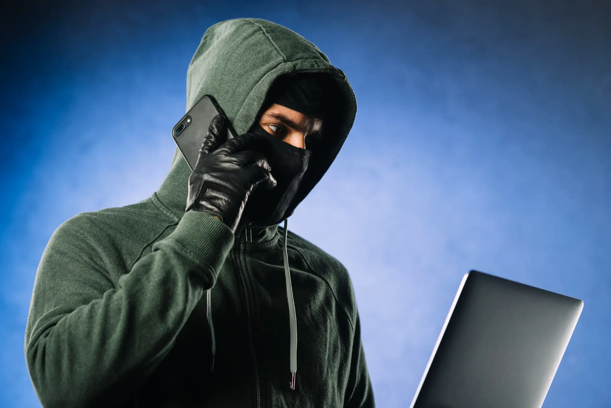 A scammer in a hoodie and mask coordinates fraud on a phone and laptop, representing TikTok's AI-driven fraudsters.