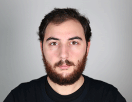 A man with dark brown hair and a full beard is captured in a close-up portrait against a plain gray background. He is wearing a black shirt and looking directly at the camera with a neutral expression. His facial features are clearly visible, including his eyes, nose, and mouth. The lighting in the image is even and illuminates his face well.
