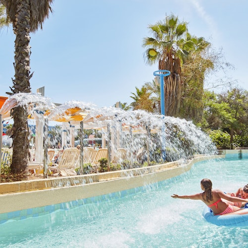 Children in swimsuits enjoy a lazy river ride on inflatables, passing through sprouting water streams under palm trees and umbrellas.
