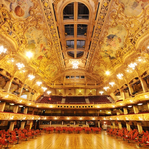 Lavishly decorated hall with a painted ceiling, chandeliers, red chairs, and an ornate balcony.