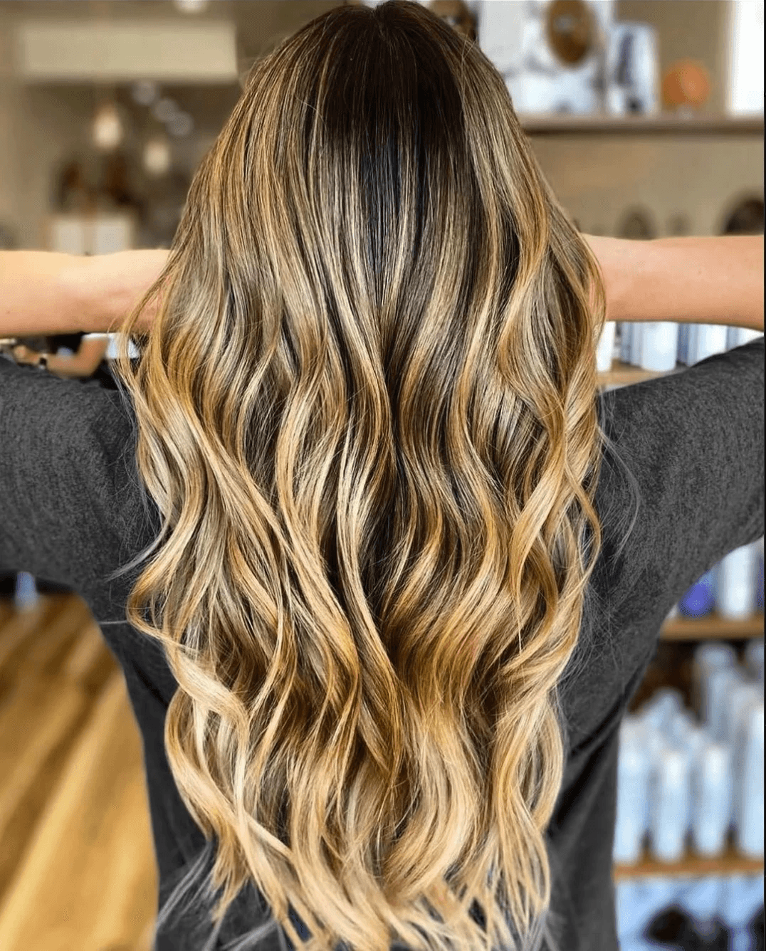 Back view of long, golden brunette hair with warm honey balayage and seamless extensions, styled in defined beachy waves for a sunlit, dimensional finish.