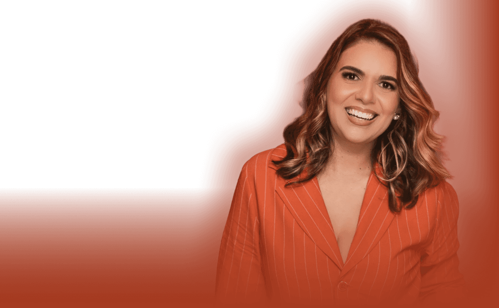 Angelina Moura, a smiling woman with wavy brown hair wearing a beige blazer and statement necklace, sitting in an elegant brown-toned setting with soft lighting.