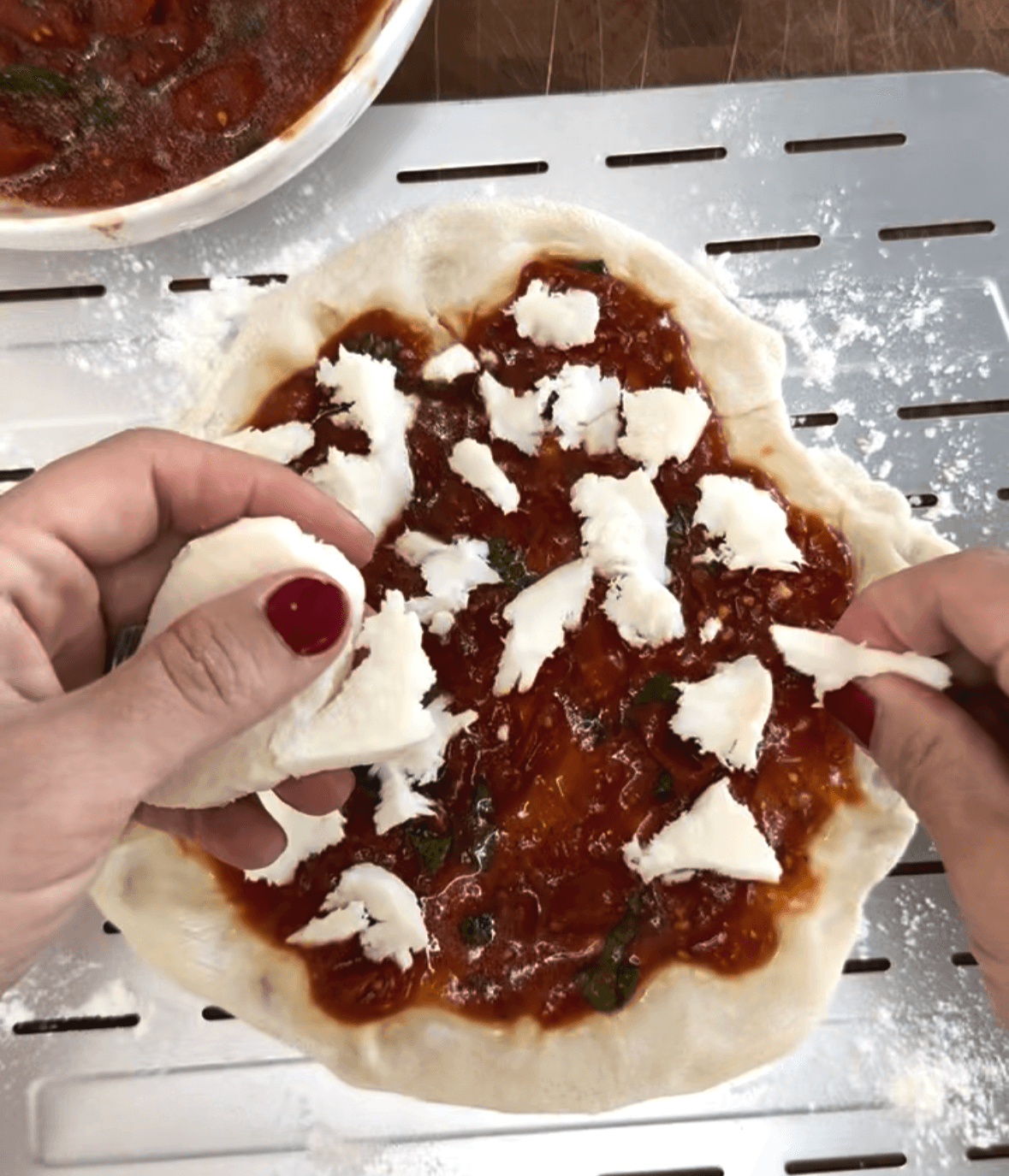 Hands adding torn mozzarella to pizza dough with tomato sauce