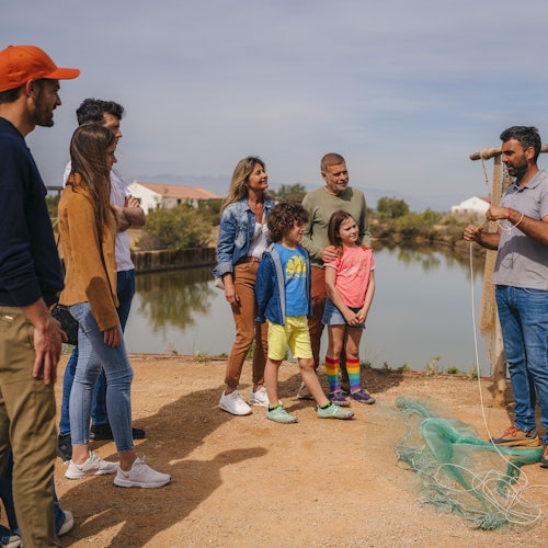 Eine Gruppe von Menschen steht an einem See und hört einem Mann zu, der etwas erklärt, das ein Fischernetz auf dem Boden betrifft.