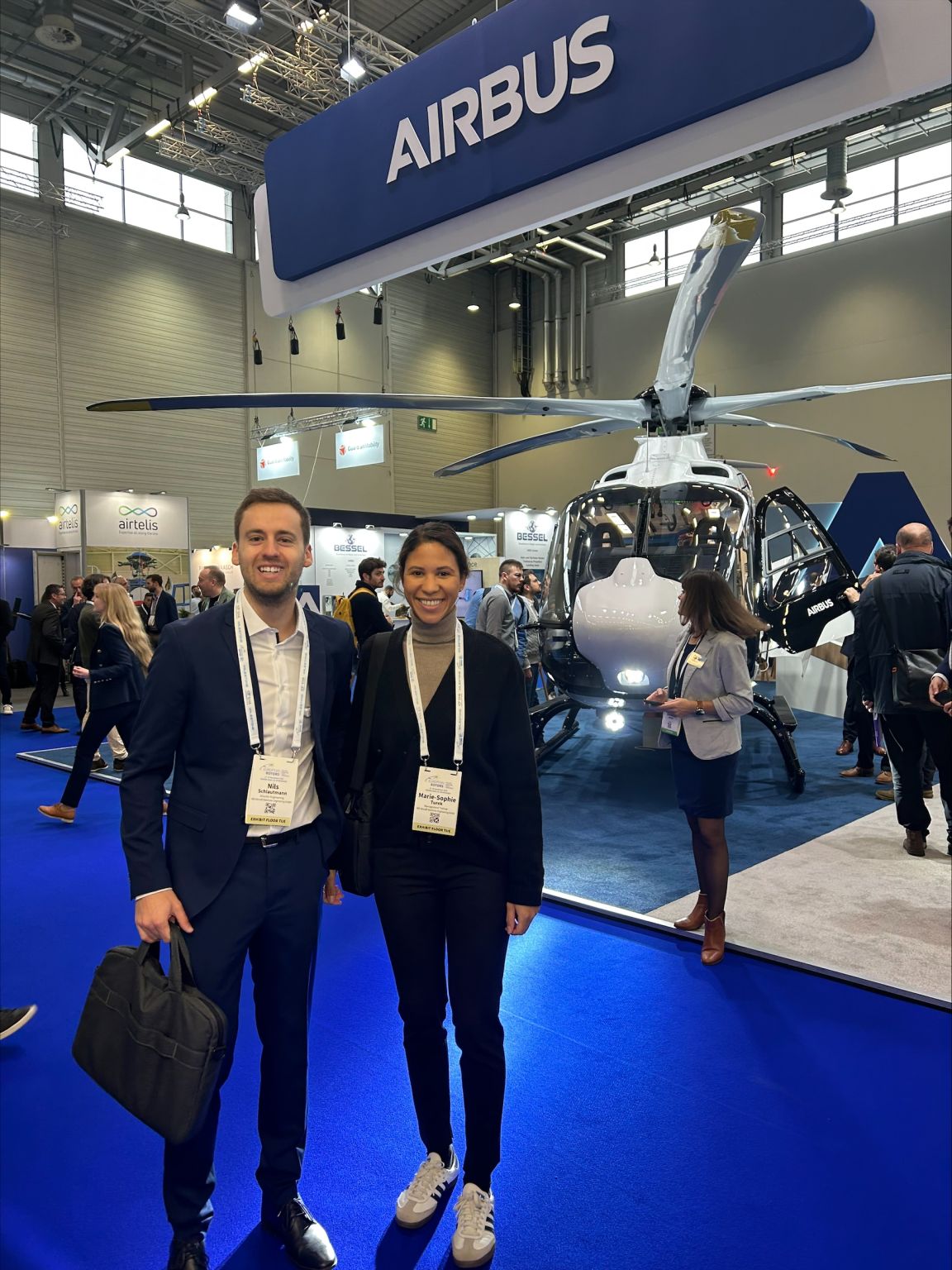 A man and a woman looking into the camera at a trade fair, with a helicopter visible in the background.