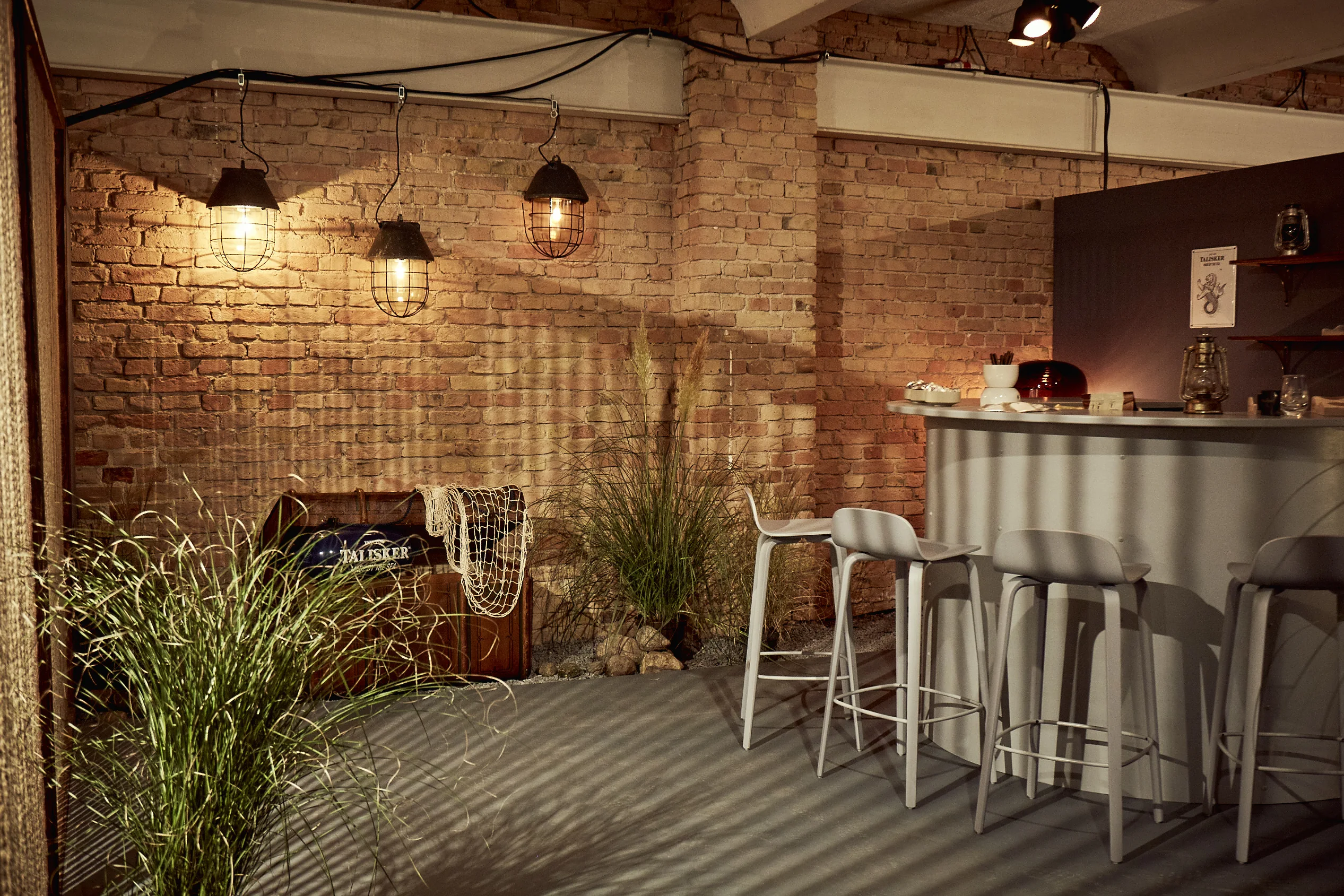 Talisker corner at DIAGEO BCB17 with a grey curved bar, high stools, caged pendant lamps, dune grass and a wooden chest against exposed brick.