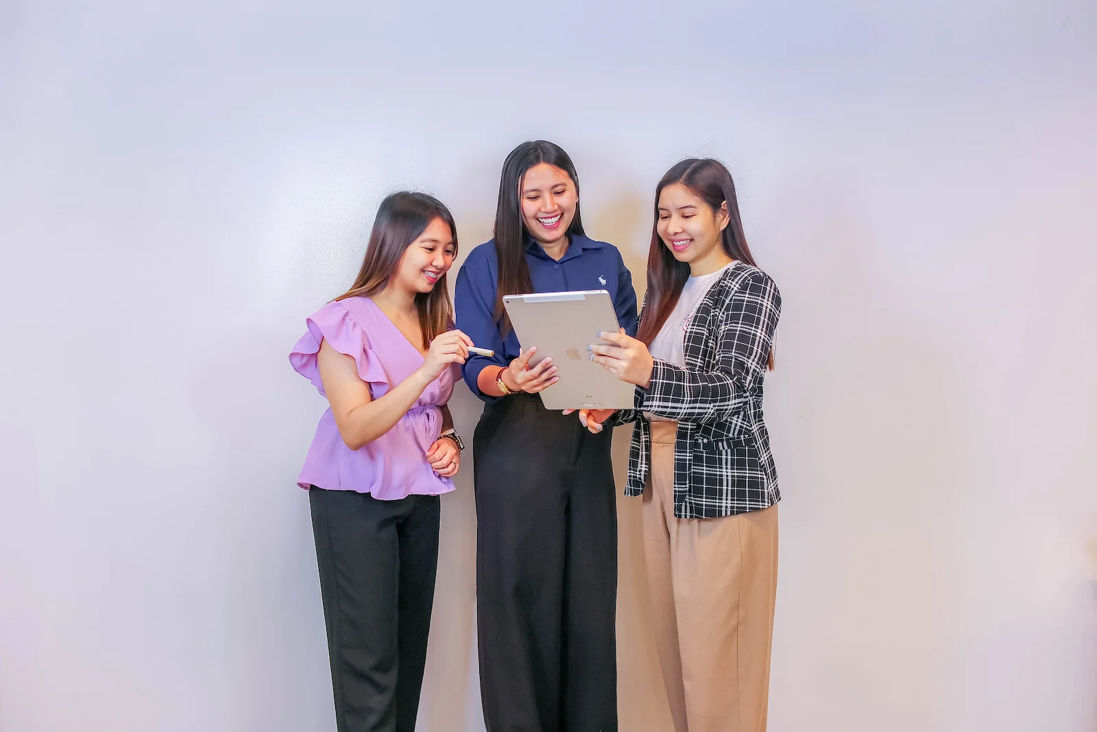 Three women stand in front of a plain white background, gathered around a clipboard, smiling as they review it together.
