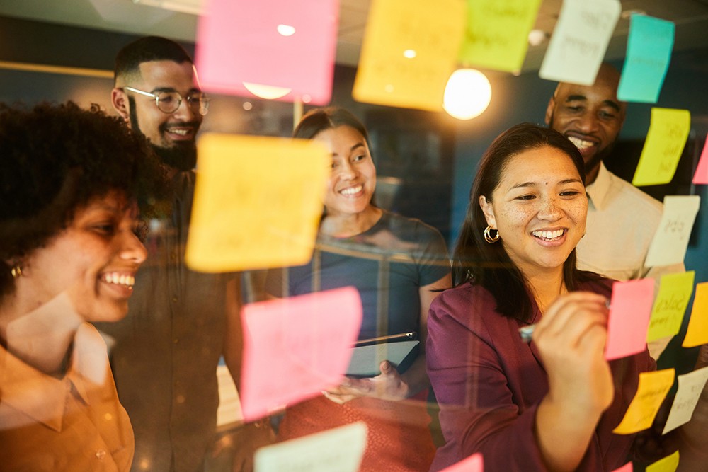 A diverse group of five colleagues smiling and writing on colourful sticky notes on a glass wall during a brainstorming session.