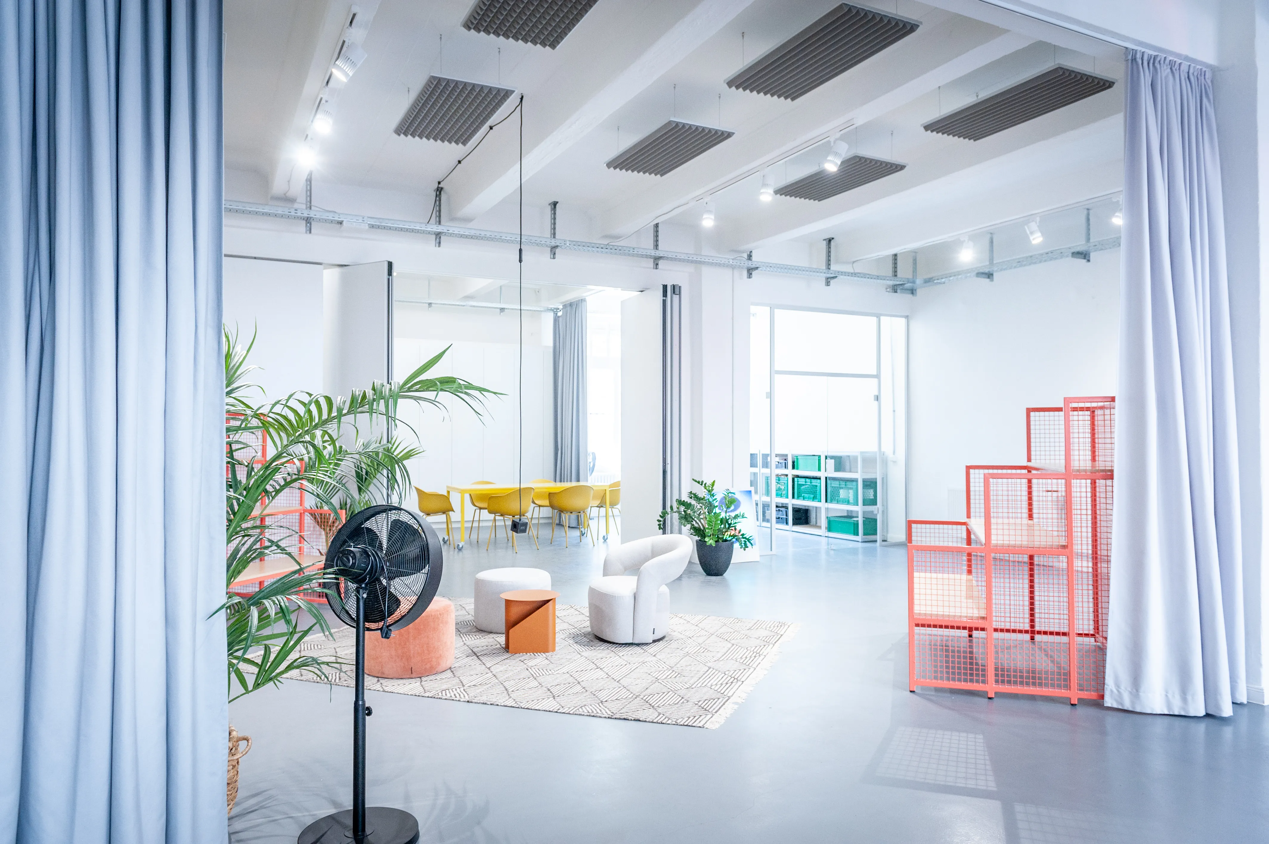 Bright lounge corner at HEY CONTENT STUDIO with white armchairs, poufs on a patterned rug, palm plants, a black fan and a coral stepped wire shelf beside light blue curtains.