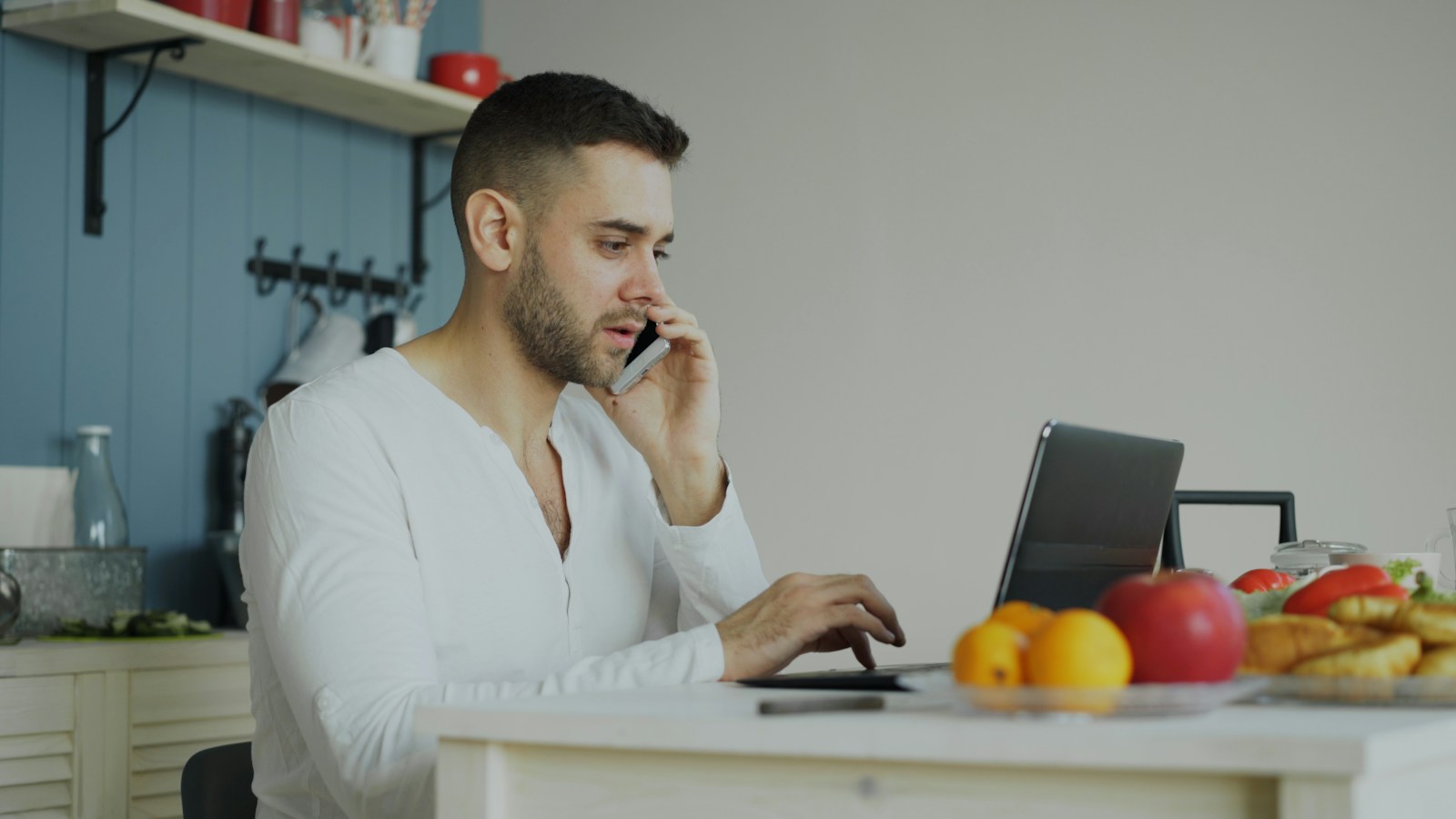 A man attends a telehealth nutrition appointment from his kitchen, talking on the phone while using a laptop near fresh fruit.