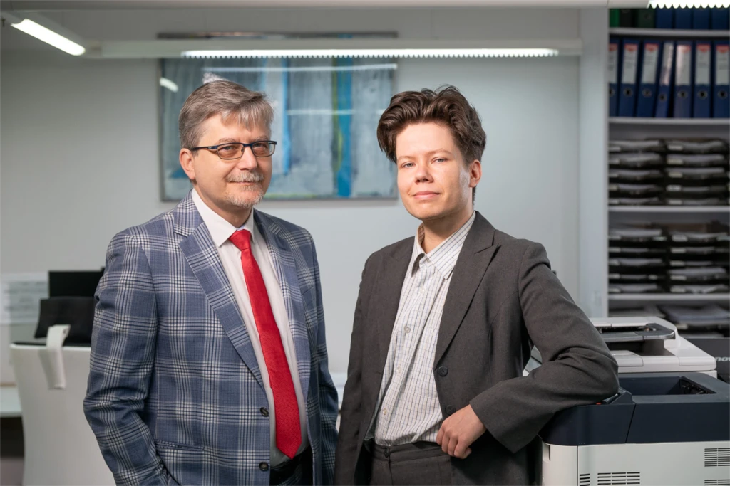 Dwóch mężczyzn w garniturach, starszy z okularami i czerwonym krawatem, młodszy uśmiechnięty, na tle biurowym.